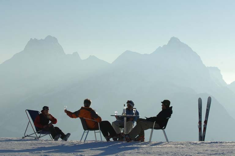 Skiers relaxing at a mountaintop table with skis planted in the snow